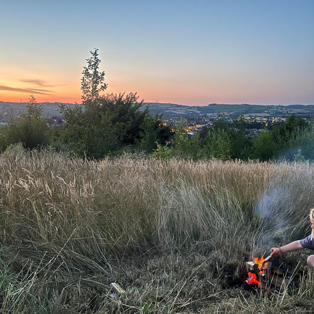 A woman crouches in a grassy field at sunset, tending a small fire. A wide landscape vista with trees, distant hills, and a glowing city skyline is visible in the background.
