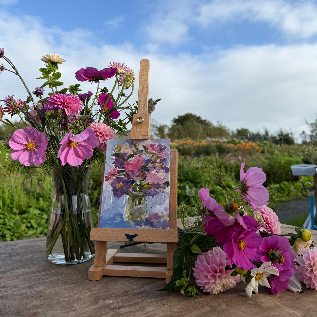 A small wooden easel holding a colorful floral painting, styled outdoors on a wooden table with pink and purple flowers in a glass vase and loose around it, against a blurred field and blue sky background.