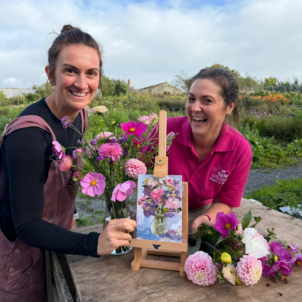Two smiling women outdoors at a table with flowers and a small painted floral artwork on an easel. One woman wears a dark apron over a black top, and the other wears a bright pink polo shirt.