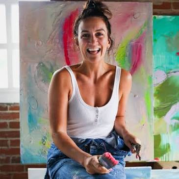 A smiling woman sitting on a stool in front of colorful abstract paintings, wearing a white ribbed tank top and blue jeans, holding paintbrushes in a studio with a brick wall background.